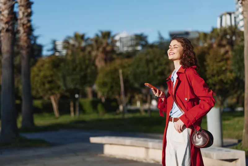 Young woman in a red coat enjoys a sunlit park moment, smiling softly as she walks with a phone in hand and a relaxed, confident stride.