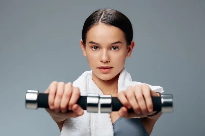 Fitness woman in a gym holds dumbbells with a towel draped over her shoulders, focused on strength training. Close-up shot highlighting effort, grip, and determined expression.