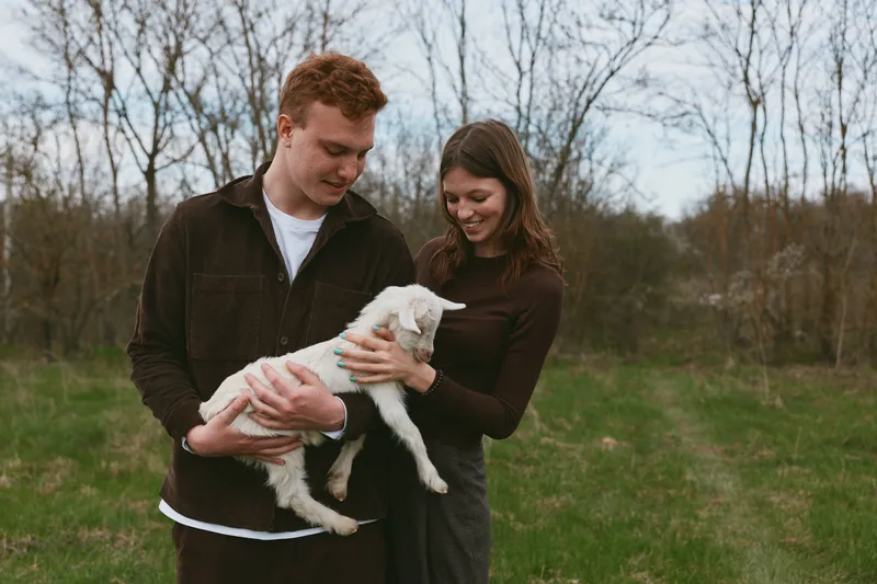 A couple outdoors gently cradle a small goat kid, smiling as they stand on a grassy path with bare trees in the background. Lifestyle moment depicting rural life, care, and a warm family bond with