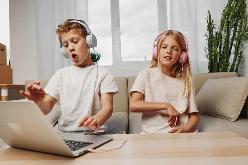 Two kids wearing headphones enjoy music and dance together in a cozy living room with natural light and green plants.