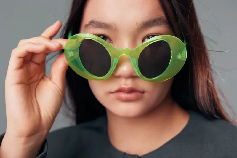 Woman wearing green round sunglasses looking at camera with serious expression closeup, fashion accessory and portrait of young female with long hair and natural makeup indoors.