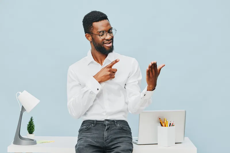 Smiling man with glasses wearing white shirt, standing at modern office desk, pointing with one hand, gesturing with the other, cheerful expression, professional workspace, light blue background, stud