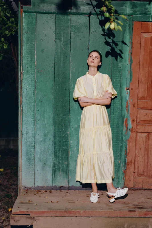 Female model in a pale yellow dress stands outdoors beside a green wall and brown door, arms crossed, looking up, conveying calm elegance and vintage style.