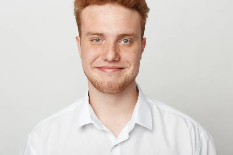 Smiling young man with red hair and beard wearing white shirt looks at camera on light background. Portrait of confident male with friendly expression and casual style.