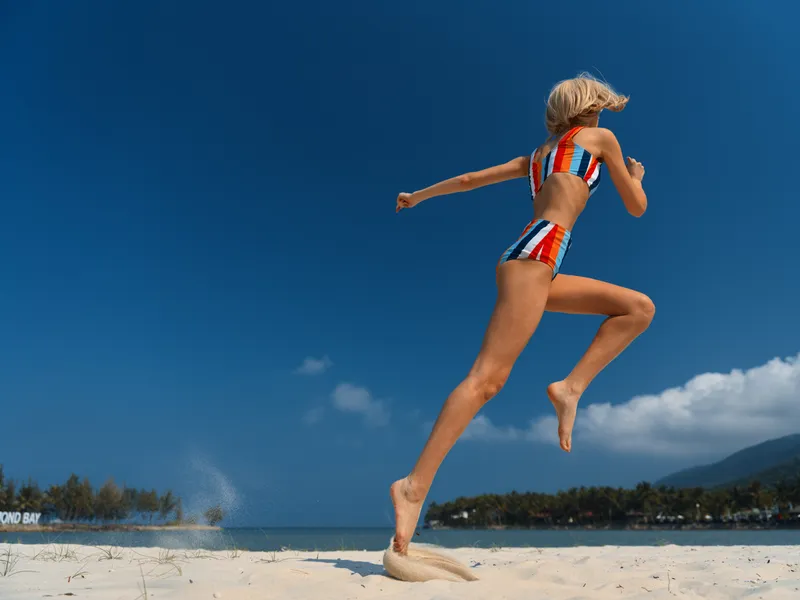 Athlete in striped bikini leaps mid air on a sunny beach, sand spray rising as feet push off, blue sky and distant waves frame energetic summer motion and freedom.