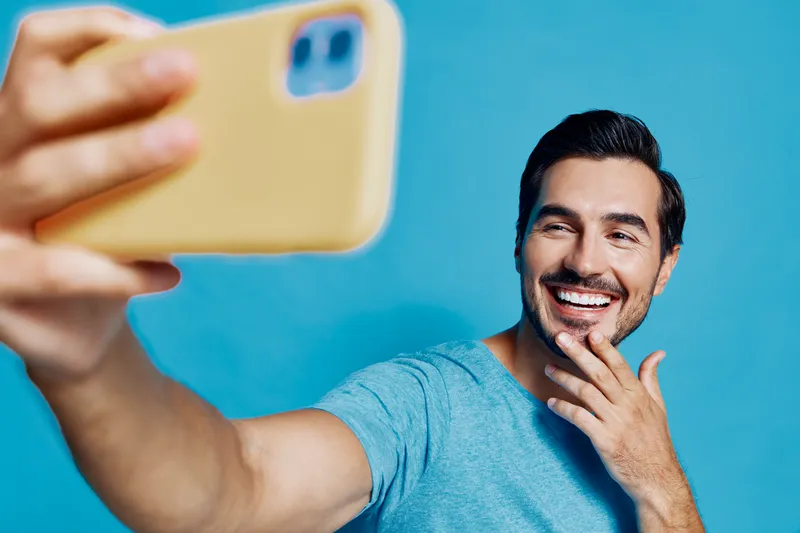 Smiling man taking selfies with smartphone, wearing casual blue t-shirt, showing cheerful expression against solid blue background. Modern lifestyle concept.