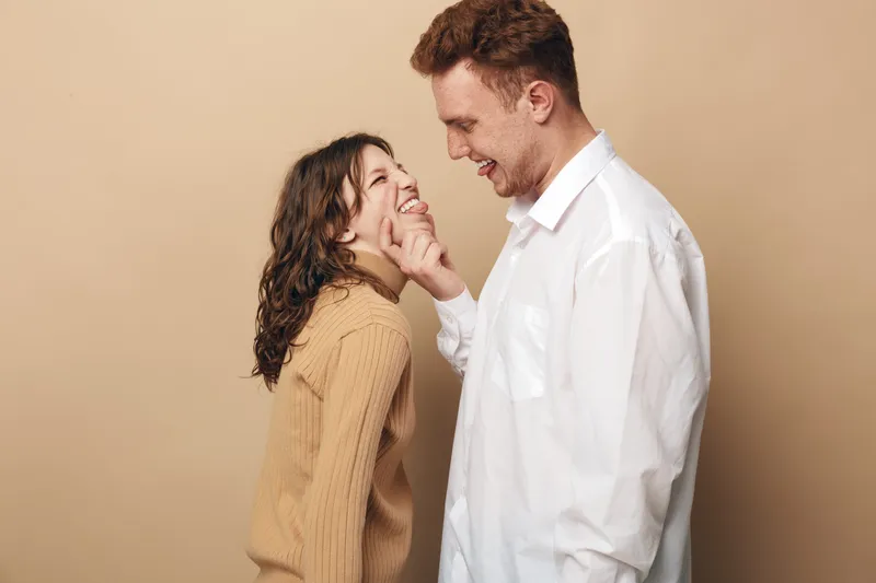 Happy young couple smiling and looking at each other, man gently touching woman’s chin. Warm beige background, casual clothing, concept of love, affection, and relationship.