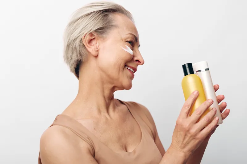 Mature woman with short grey hair smiling and holding two skincare bottles while applying cream on her cheek. Concept of beauty routine, healthy skin, anti aging and self care.
