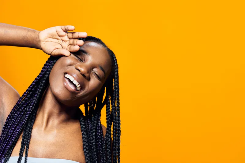 Joyful young Black woman with braided hair smiling against a vibrant yellow background, conveying happiness and positivity in a playful manner
