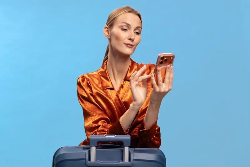 Young woman in orange satin blouse holds smartphone and looks at screen while standing behind blue suitcase. Concept of travel planning, technology use, and modern lifestyle with vibrant colors.