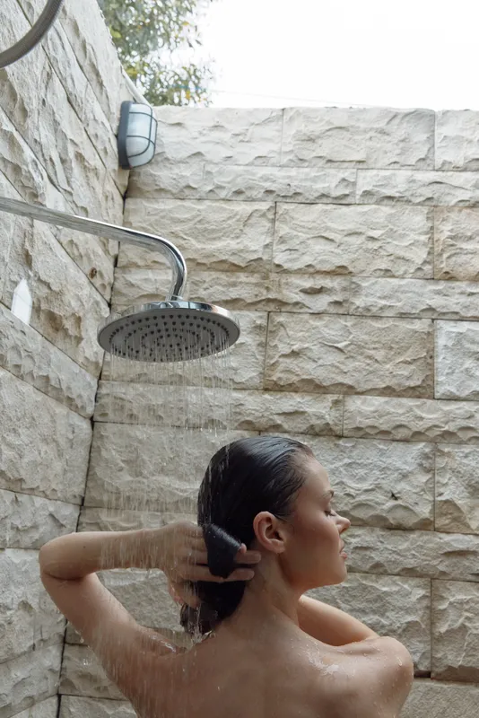 woman showering outdoors under rain shower head, natural stone wall background, relaxed posture, fresh skin cleansing, wellness concept.