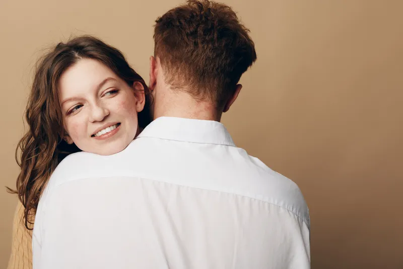 Smiling woman with brown hair embraces man wearing white shirt on beige background. Intimate moment of affection and connection between couple in casual clothing. Warm, soft lighting.
