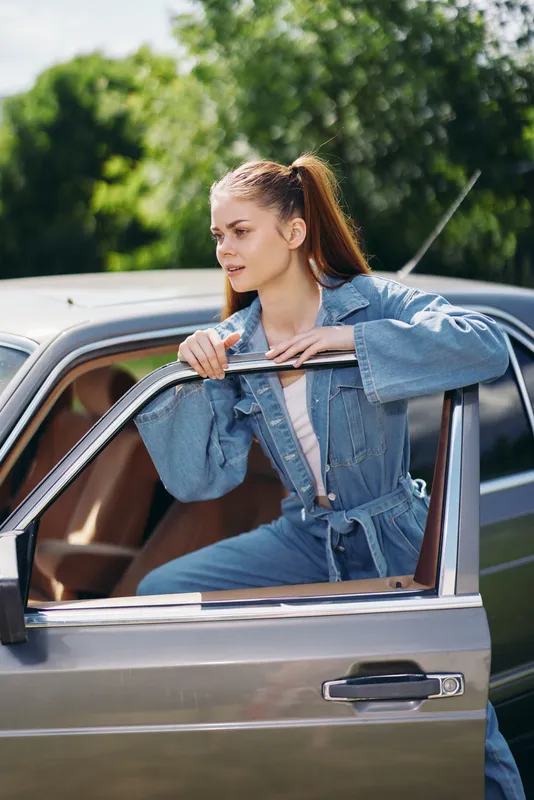 Stylish young woman in denim jumpsuit posing by vintage car outdoors, showcasing a modern yet timeless fashion sense, embodying confidence and style.