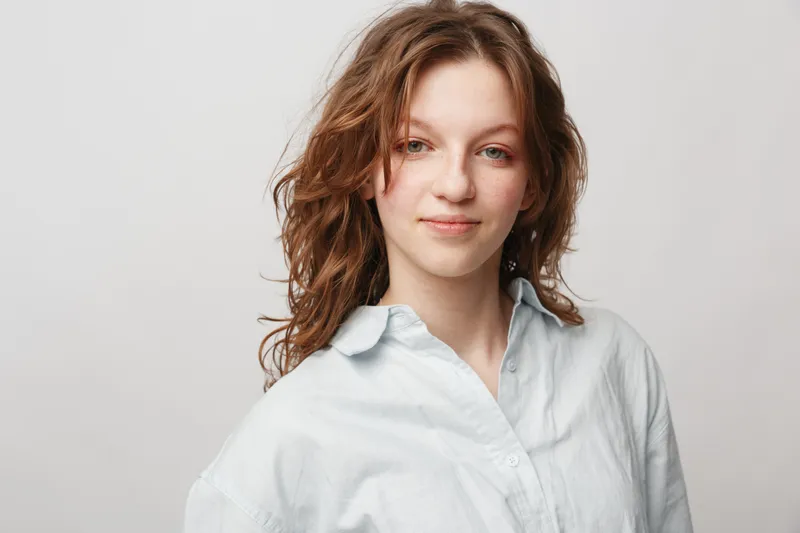 Young woman with wavy hair wearing white shirt smiles gently against plain background. Portrait of confident female with natural look and casual style in studio lighting.