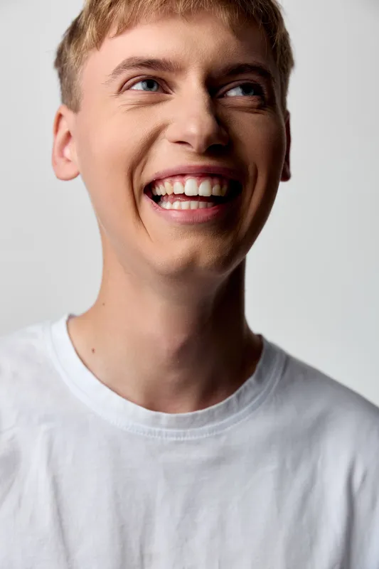 Smiling young man in a white tee with bright expression, open laughter, and relaxed posture, conveying happiness, optimism, and friendly energy in a studio setting