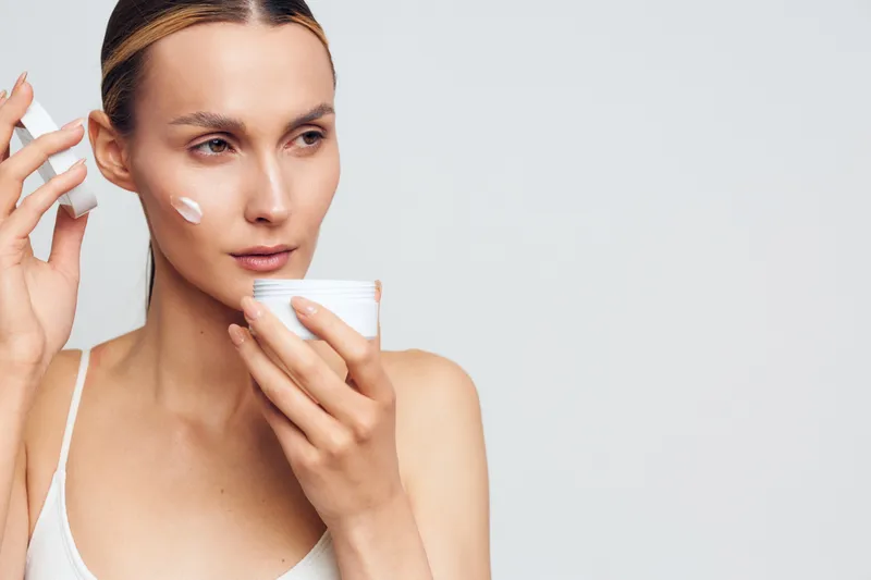 Young woman with blonde hair applies moisturizing cream on her face while holding a jar in a bright studio, showing skincare routine and natural beauty care.