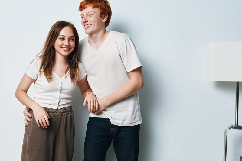 Young couple smiling and standing close together with casual clothing against light background. Happy man and woman showing friendship and relaxed lifestyle indoors.