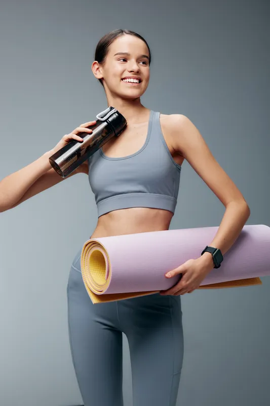 Woman in athletic wear holds a water bottle and a rolled yoga mat, ready for a workout and fitness routine in a bright studio. Her confident stance reflects motivation and health during training.