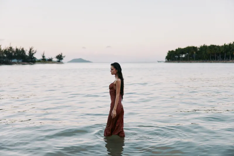 A lone woman stands waist-deep in calm coastal water, wearing a flowing red dress, as the warm sunset light shapes her silhouette and mood.