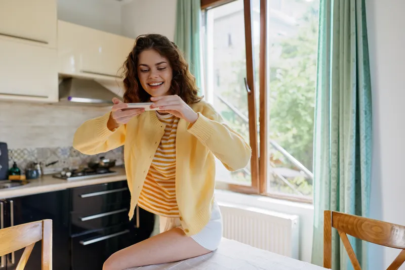 Smiling woman in a yellow cardigan captures a moment on a smartphone in a bright kitchen, expressing casual home life with daylight and a cozy, welcoming mood.