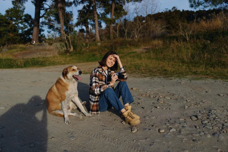 A woman and dog sit outdoors on gravel, casual plaid shirt and boots, enjoying nature and a sunny day with trees in the background, peaceful moment of companionship