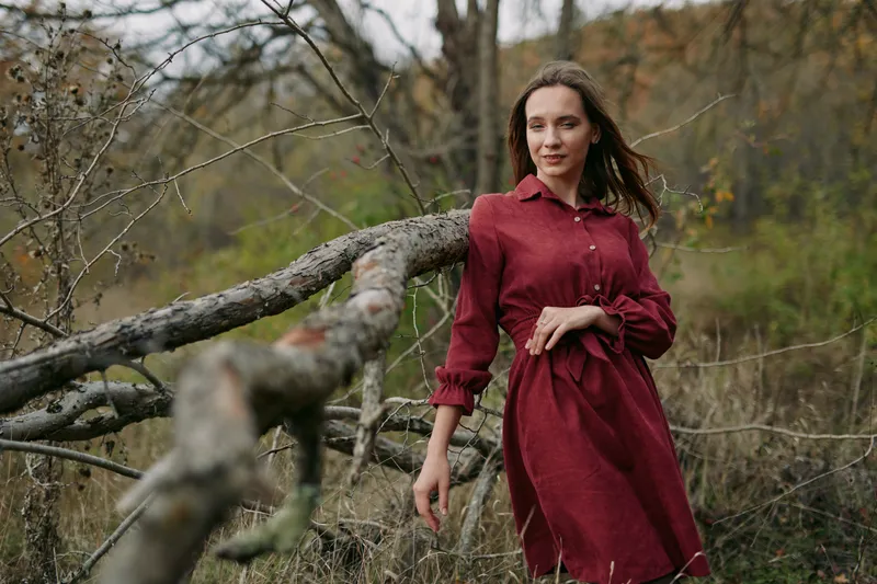 Portrait of a woman in a red dress standing beside a weathered branch, conveying authentic mood and natural pose in a calm rural landscape.