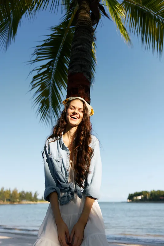 Portrait of a smiling young woman standing beside a tall palm on a sunny beach, wearing a denim shirt and light dress, exuding carefree vacation charm.