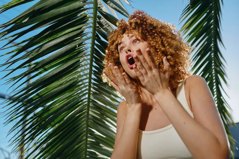 curly woman shouting outdoors with palm leaves background, natural light, casual white tank top, expressive emotion, sunny summer day, vibrant green foliage