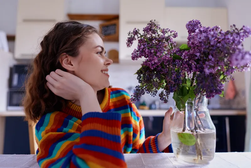 A smiling woman in a colorful striped sweater sits at a cozy kitchen table, admiring a vase of purple flowers. A warm, candid moment of home life and comfort.