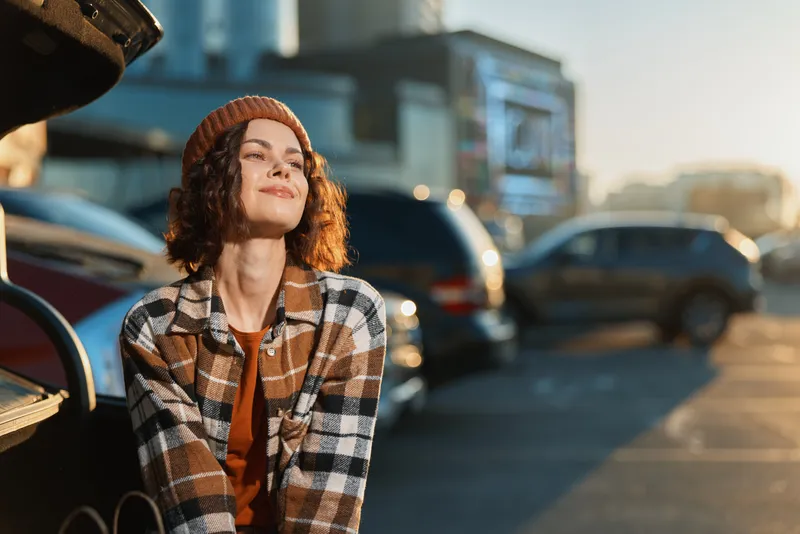 Woman in beanie and plaid jacket sits by open car trunk in parking lot, sunlight on her face, candid smile and golden hour glow conveying authenticity, mindful living and emotional storytelling