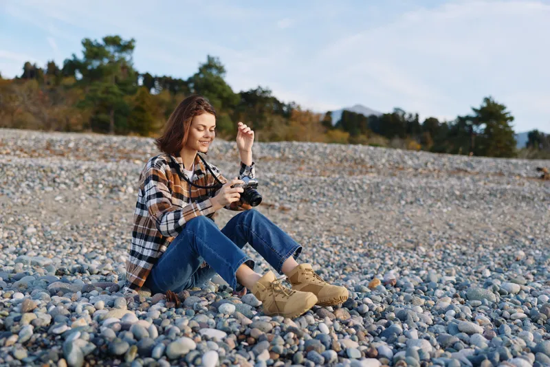 Woman sitting on a pebble beach in casual outdoor attire, focused on a smartphone, surrounded by tranquil nature, rocky shore and open sky, conveying relaxed connectivity.