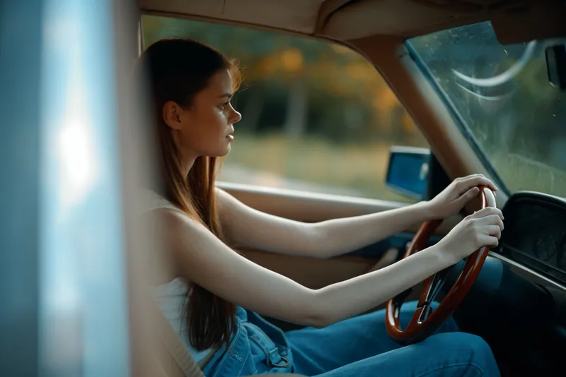 A young woman with long hair driving an old car, showcasing confidence and adventure in a serene outdoor setting, ideal for travel and lifestyle concepts.