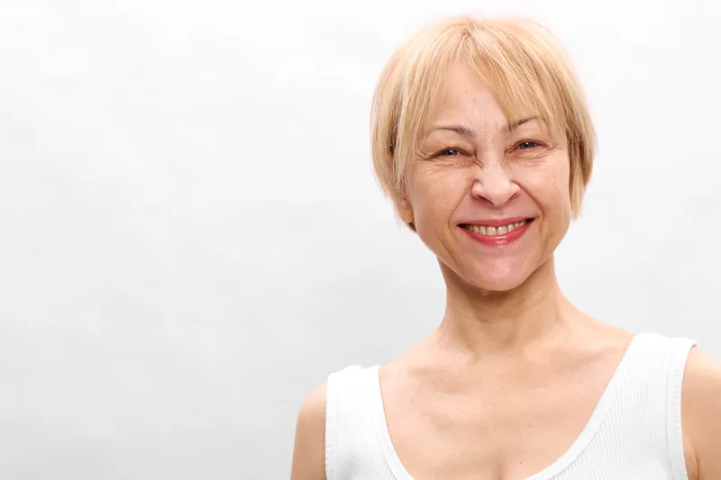 Woman portrait smiling elderly blonde hair wearing light clothing on white background, close framing, concept of skincare wellness and self care, soft studio lighting creates calm warmth
