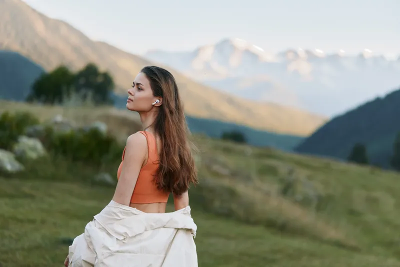 A woman stands in a golden landscape, gazing toward distant mountains. She wears an orange top and light jacket, embodying calm, exploration, and outdoor freedom.