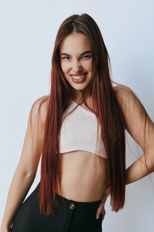Young woman with long straight hair smiling confidently posing in casual white crop top and black pants against plain light background with hands on hips and friendly expression.