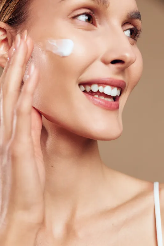 Smiling woman applies face cream on cheek with fingers closeup healthy skin natural beauty skincare routine isolated on beige background with happy expression