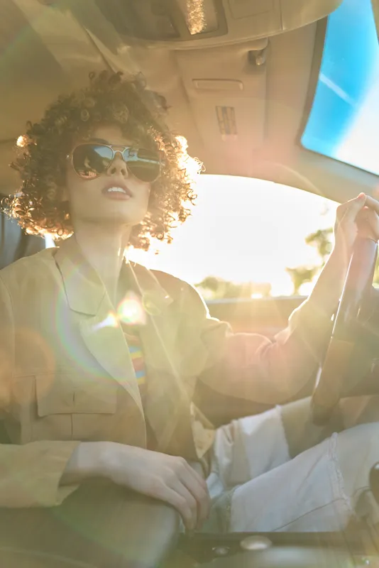 A woman with curly hair wearing sunglasses drives a car in warm retro light, capturing a candid lifestyle moment and themes of freedom and road trips. Her relaxed pose and sunlit interior evoke a