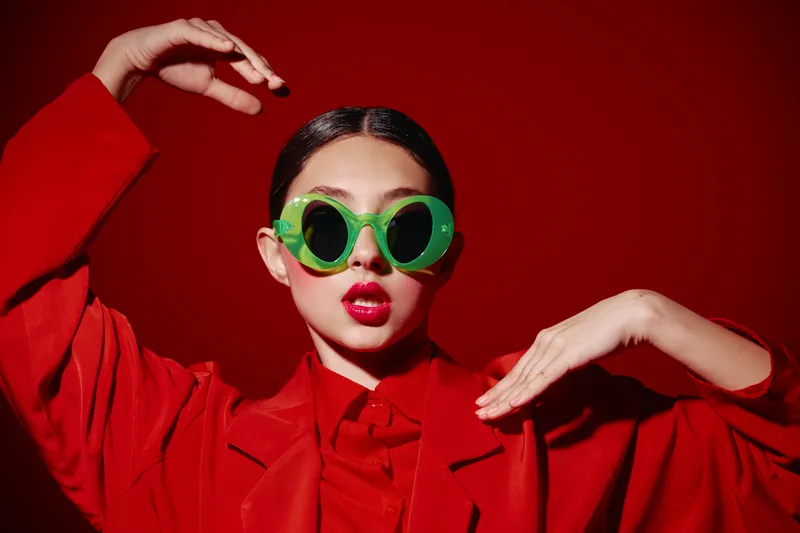 Stylish woman wearing bright green sunglasses and an oversized red suit makes artistic hand gestures against a matching red background. Bold fashion and beauty concept.