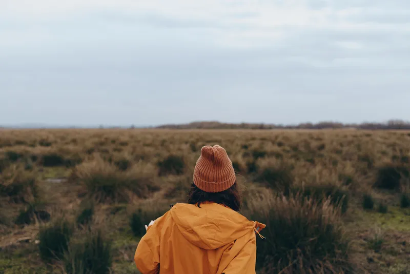 Woman lifestyle outdoors in nature, seen from behind in a field grassland, wearing warm jacket and hat, watching horizon on an overcast day, calm travel mood.