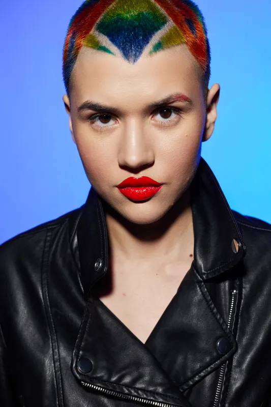 Young woman with colorful rainbow hair and bright red lipstick wearing a black leather jacket against blue background, showcasing bold edgy style and confident attitude.