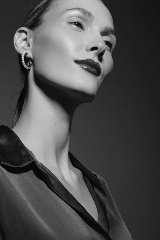 Elegant white woman with fair skin poses confidently in black satin blouse. Studio portrait in black and white with dramatic lighting highlights her glossy lips and strong jawline.