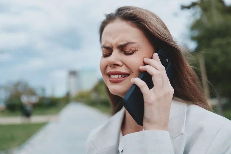 Woman stressed on phone call showing emotional distress and frustration outdoors in casual style KEYWORDS: woman stressed phone call emotional distress frustration outdoor casual communication anxiety