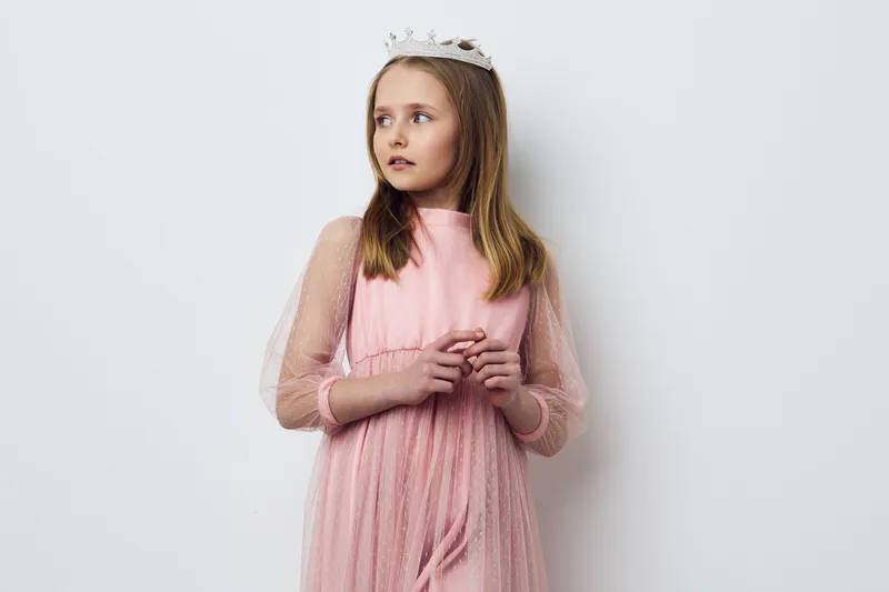 A young girl in a pink tulle dress with sheer sleeves and a crown, looking pensive against a neutral background, capturing a whimsical and dreamy atmosphere