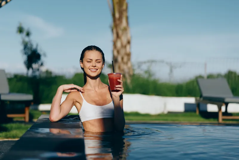 Young woman relaxes in a sunny pool, holding a juice cup and smiling, wearing a white bikini top, with palm trees, lounge chairs, and a clear blue sky forming a vibrant outdoor backdrop