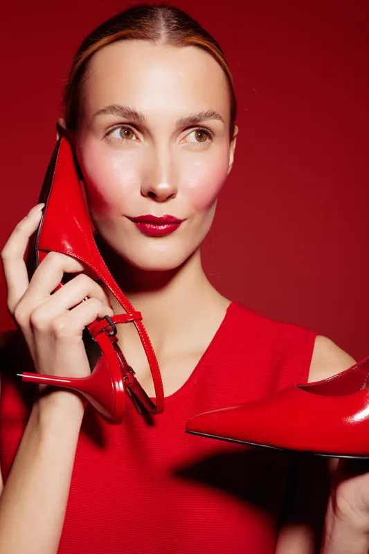 Woman in red dress holds red high heel shoes near face against red background. Fashion concept with stylish accessories and bold color contrast for modern elegance and glamour.
