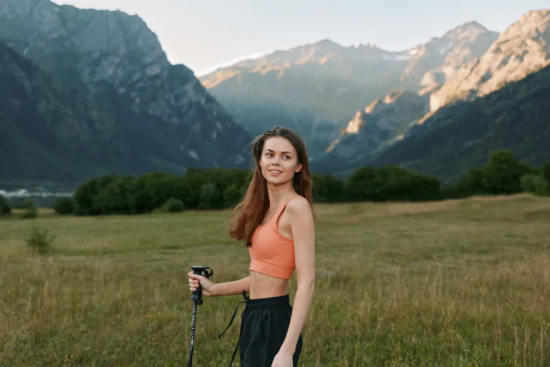 A young woman hiker stands in a mountain meadow with trekking poles. She wears an orange sports top and black shorts, smiling as the scenic valley and rugged peaks frame her adventure.