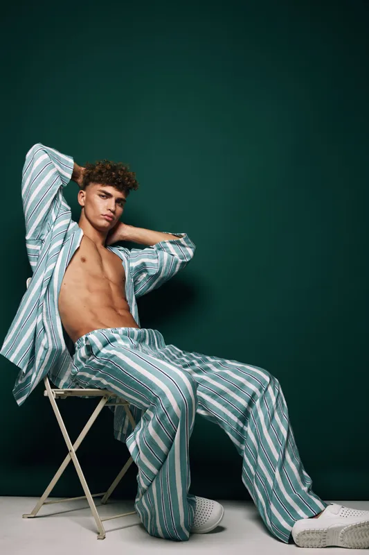 Young man with curly hair wearing striped pajamas sitting on chair with hands behind head against dark green background in studio. Relaxed male model posing with casual sleepwear and white shoes.