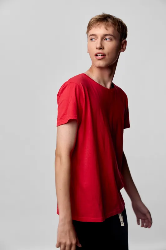 Young man in a red t shirt stands against a neutral background, looking to the side, showcasing casual fashion and a relaxed studio vibe for lifestyle and apparel projects