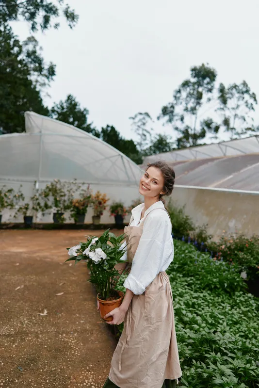 Young woman in an apron smiling while holding a potted plant in a greenhouse, showcasing joy and connection to nature Seasonal gardening theme
