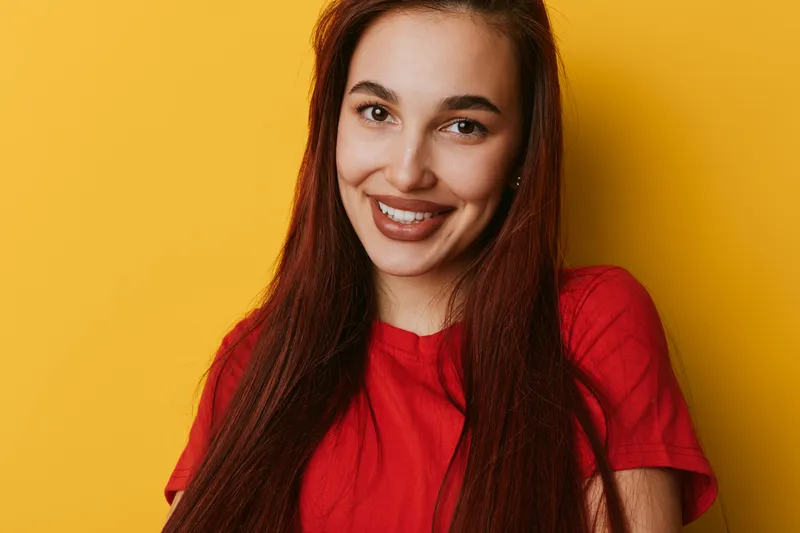 Young woman with long red hair wearing red shirt smiling and posing against yellow background. Portrait of happy female with natural makeup and confident expression in studio.